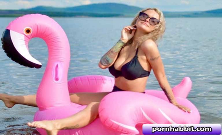 Woman in a black bikini lounging on a large pink inflatable flamingo in calm water, wearing sunglasses and posing for the camera.