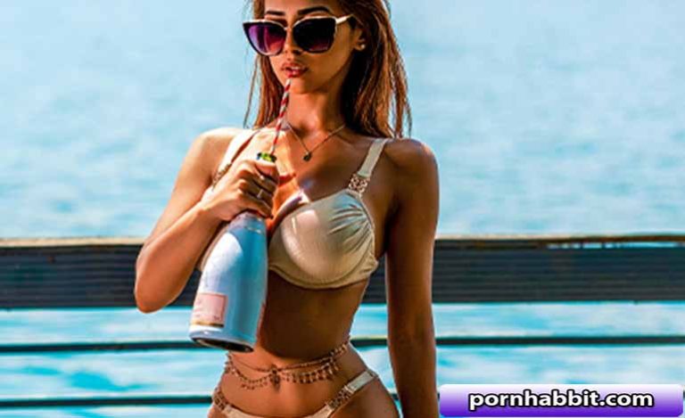 Woman in a beige bikini sipping from a champagne bottle on a sunny seaside pier, water in the background.