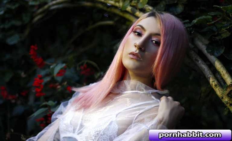 Model with pink hair posing among greenery in a sheer white lace top, looking softly at the camera as red flowers and vines frame her portrait.